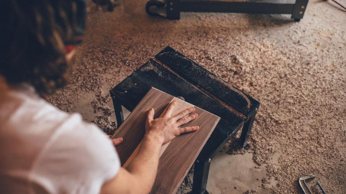 Persona lijando a mano una tabla de madera sobre un banco de trabajo con serrín alrededor